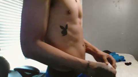 Media: Video of a shirtless man with a muscular build, light skin, and a black butterfly tattoo on his left ribcage. He wears blue shorts and stands in a dimly lit room with white blinds partially covering a window.