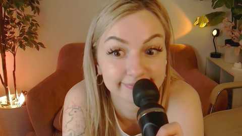 Media: Video of a fair-skinned, blonde-haired woman with long hair, smiling while holding a microphone. She has a tattoo on her right arm and is seated in a cozy, dimly lit room with warm lighting and greenery.