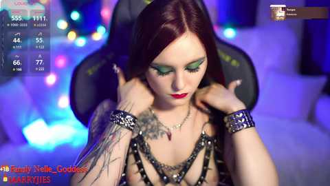 Media: Video of a pale-skinned woman with red hair, wearing bold green eyeshadow and a black spiked choker, seated on a gaming chair in a dimly lit room.