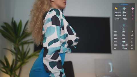 Media: Video of a curvy woman with long blonde hair, wearing a patterned top and high-waisted blue pants, standing in a modern room with a flat-screen TV, a potted plant, and a digital interface displaying weather and temperature data.