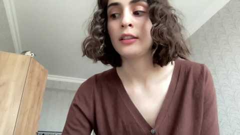 Media: Video of a young person with curly dark hair, wearing a brown V-neck cardigan, standing in a modern room with light grey patterned wallpaper and wooden furniture.