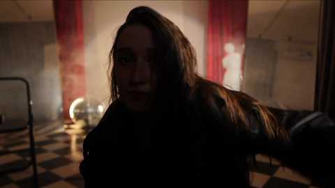 Media: Video of a young woman with long brown hair, wearing a black leather jacket, seated in a dimly lit room with red curtains, a checkered floor, and a metal chair in the background.