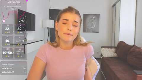Media: Video of a young Caucasian woman with light brown hair, wearing a pink t-shirt, sitting in a modern living room with a TV and sofa in the background.