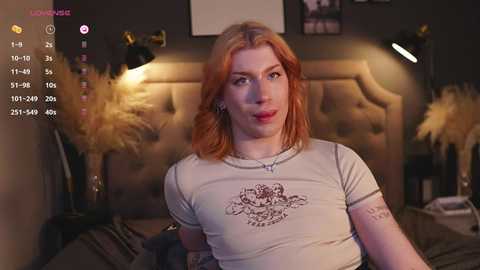 Media: Video of a fair-skinned, red-haired woman with a tattooed arm, wearing a gray crop top with a detailed design, sitting on a bed in a dimly-lit bedroom.