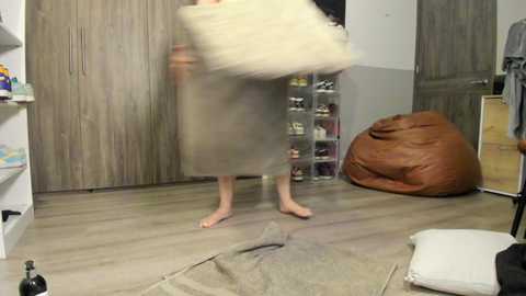 A video of a blurred person in a grey dress, barefoot, holding a beige cushion. Wooden door, shoe rack, and brown beanbag chair in a modern, minimalist room with light wooden flooring.