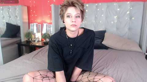 Media: Video of a pale-skinned, androgynous person with short, spiky, blonde hair, wearing a black T-shirt and fishnet stockings, sitting on a bed with gray bedding and a tufted headboard.