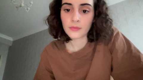 Media: Video of a young woman with fair skin, curly brown hair, and a neutral expression, wearing a brown sweater, indoors against a light gray wall.