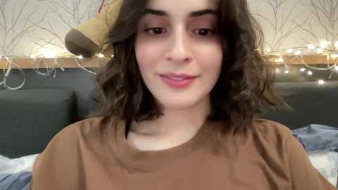 Media: Video of a young woman with wavy brown hair, fair skin, and a relaxed expression, wearing a brown top, seated on a gray couch in a cozy room with string lights and a stuffed animal in the background.