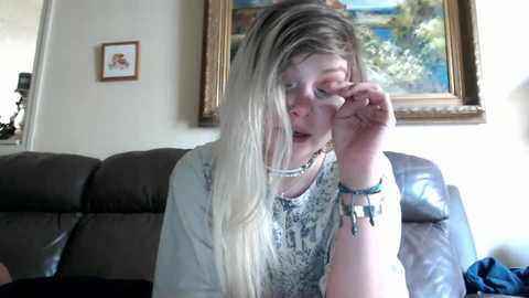 Media: Video of a young woman with long, straight, blonde hair, wearing a light blue patterned top and a pearl necklace, adjusting her glasses, seated on a brown leather couch.