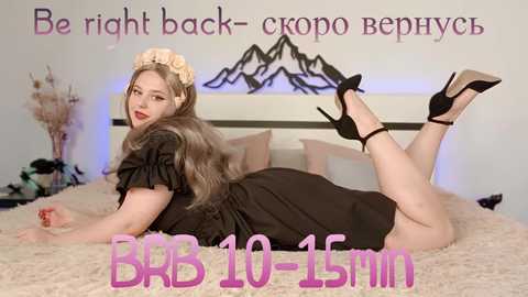 Media: Video of a plus-size woman with light skin, long brown hair, and wearing a black dress and black high heels, lying on a beige bedspread in a bedroom.