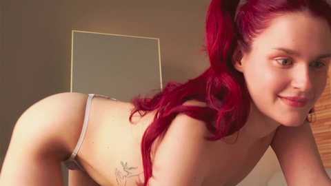 Video of a topless woman with vibrant red hair styled in pigtails, wearing a thong, posing in a suggestive manner.