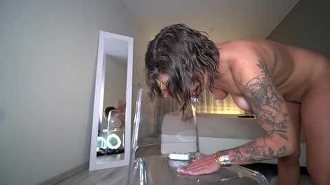 Video of a naked, tattooed woman with wet, shoulder-length brown hair, leaning over a clear acrylic chair, using a smartphone to take a selfie in a modern, minimalistic bedroom.