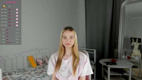 Media: Video of a young, fair-skinned, blonde girl with straight hair, wearing a white t-shirt, sitting on a bed in a sparsely furnished, grey-toned room with a calendar and a mirror.