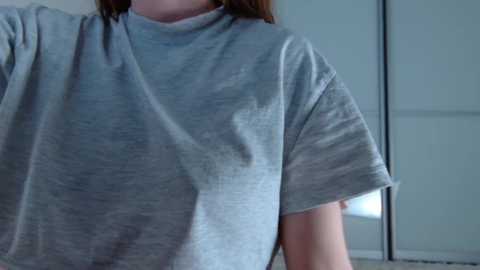 A video of a light-skinned person wearing a loose, short-sleeved, light grey t-shirt. The background shows a blurred, modern interior with glass doors and soft lighting. The shirt's texture appears soft and slightly wrinkled.