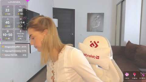 Media: A video shows a blonde woman in a beige top playing a video game on a gaming chair with a white headrest. The room has a modern, minimalist design with white walls and dark furniture.