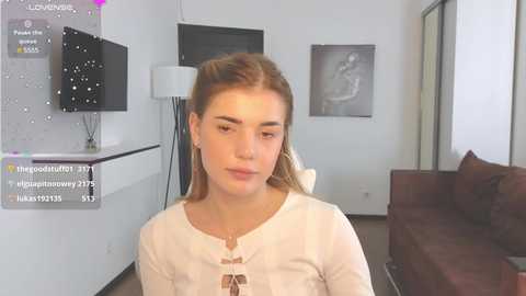 Video of a young, light-skinned woman with long, light brown hair, wearing a white blouse, seated in a modern, minimalist living room with a TV and abstract art on the walls.