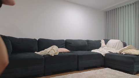 A video of a dark grey L-shaped sectional sofa in a modern living room, with white pillows and blankets strewn about, and light grey curtains covering a window.