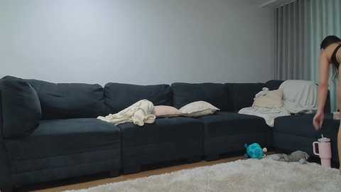 Video of a dark gray sectional sofa in a minimalist living room, with a person in a sleeveless top standing beside it, holding a pink container.