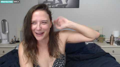 Media: Video of a smiling, fair-skinned woman with long, wavy brown hair, wearing a black and white patterned bra, lying on a navy blue bed in a modern bedroom with white walls and a bedside table.