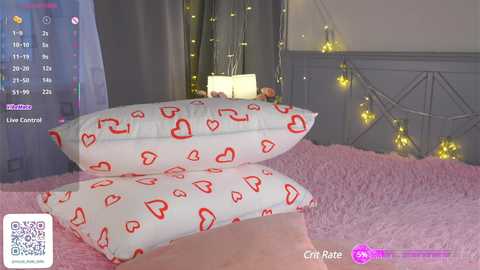 Video of a cozy bedroom with a pink plush bedspread, white pillows with red heart patterns, a grey wall, and fairy lights.