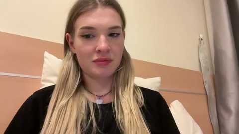 Video of a young Caucasian woman with straight, long blonde hair, wearing a black top, sitting in a hospital bed with white pillows and beige walls.