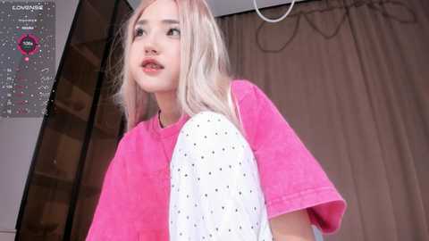 Media: Video of a young woman with light skin and platinum blonde hair, wearing a pink t-shirt with a white polka dot design, standing in a modern room with beige curtains and a white ceiling light.