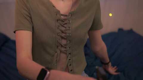 Video of a fair-skinned woman in a green, lace-up top, sitting on a bed with dark blue sheets. The background is blurred with warm lighting.
