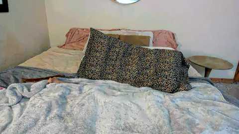 Media: Video of a cozy, unmade bed with a leopard-print pillow and a soft, textured gray blanket. A wooden side table with a round top sits nearby, and the room's walls are off-white.