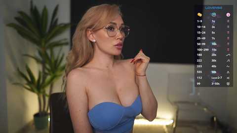 Media: Video of a blonde woman with large breasts, wearing a strapless blue dress, and glasses, holding a nail file, indoors, background includes a potted plant and dimly lit room.