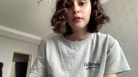 Media: Video of a young girl with wavy brown hair, wearing a light gray \"California\" t-shirt, standing in a room with white walls and a floral patterned wallpaper.