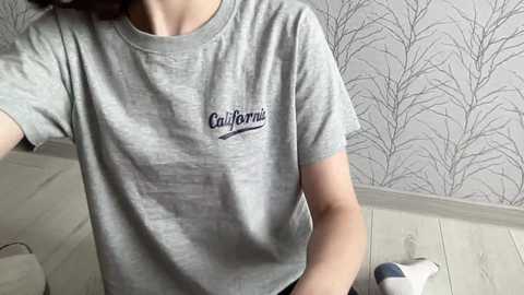 Media: Video of a woman wearing a gray t-shirt with \"California\" logo, standing in a room with white wooden floor and gray leaf-patterned wallpaper.