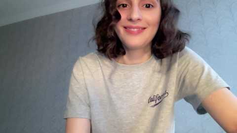 Media: Video of a smiling young girl with wavy brown hair, wearing a gray t-shirt with a small logo, standing against a textured light blue wall.