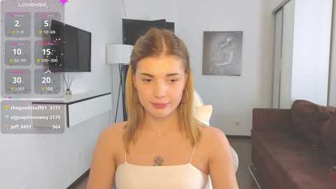 Media: A video of a young woman with fair skin and straight blonde hair, wearing a white spaghetti-strap top, sitting in a modern living room with a black TV and white floor lamp.