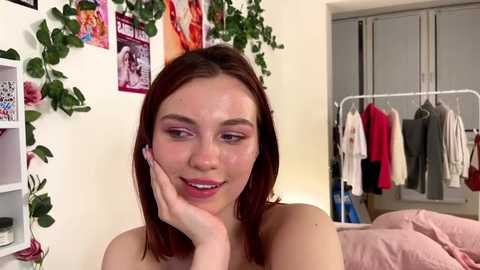 Media: Video of a young woman with fair skin, straight brown hair, and light makeup, touching her cheek. Background includes a white wall with posters, a closet, and a pink bed.