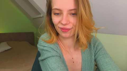 A video of a fair-skinned woman with shoulder-length, wavy, blonde hair, smiling slightly, wearing a teal cardigan, in a room with light green walls and a bed with beige bedding in the background.