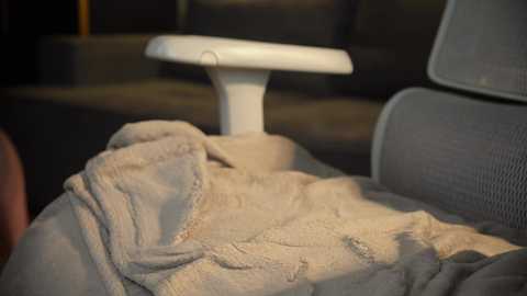 Media: Video of a beige blanket draped over a white office chair, with a blurred background of dark furniture and dim lighting. The blanket has a soft, textured fabric.
