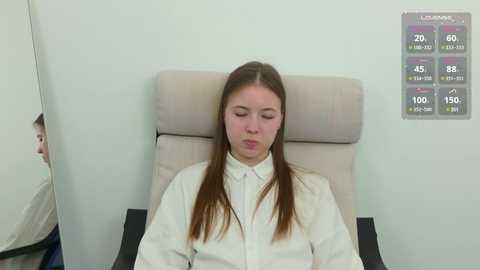 A video of a young Caucasian woman with long brown hair, wearing a white lab coat, sitting on a beige chair, with a medical oxygen saturation monitor displaying her oxygen levels.