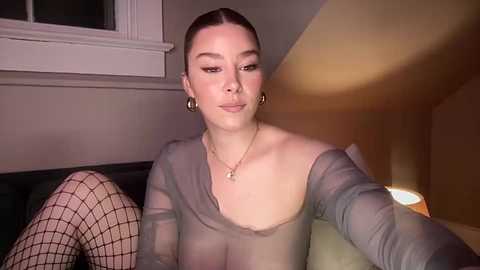 Media: Video of a light-skinned woman with straight brown hair, wearing a sheer gray top and black fishnet stockings, sitting on a bed, looking into the camera.