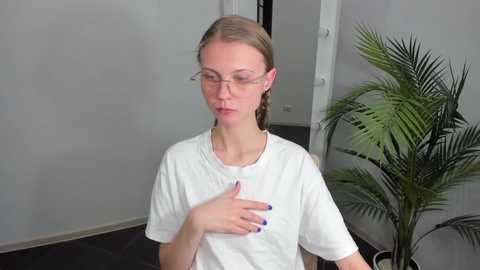 Media: Video of a young Caucasian woman with fair skin and straight brown hair, wearing glasses, a white T-shirt, and blue nail polish. She is touching her chest in a domestic setting with a potted plant and white walls.
