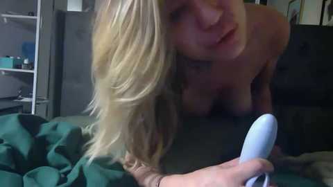 A candid video shows a topless blonde woman with long hair, lying on a bed with green sheets, using a white vibrator.