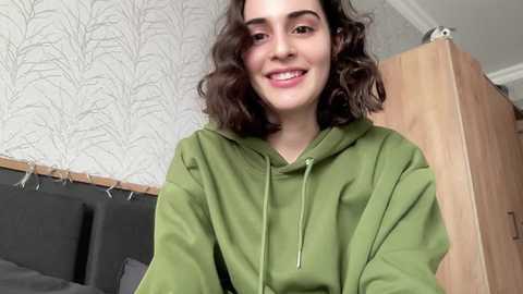 Media: Video of a young woman with curly dark hair and fair skin, wearing a green hoodie, smiling in a bedroom with white patterned wallpaper and a wooden door.