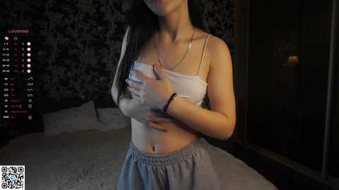 Media: Video of a slim, light-skinned woman with long black hair, wearing a white tank top and grey pants, covering her chest with her hands, in a dimly-lit bedroom with black floral wallpaper, a bed, and a lit candle.