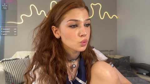 Media: Video of a young woman with wavy brown hair, fair skin, and a neutral expression, wearing a dark blouse and several necklaces, in a modern living room with gray walls and a heart-shaped light display.