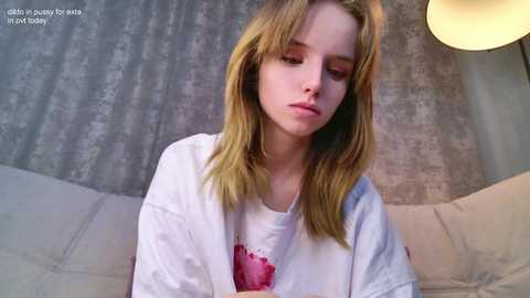 Media: Video of a young, fair-skinned girl with shoulder-length blonde hair, wearing a white t-shirt, sitting on a beige couch against a grey, patterned wall, looking somber.