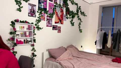 Media: Video of a pink-themed bedroom with a white bed, pink bedding, and green ivy hanging from the wall. Framed photos, a shelf, and a hanging coat rack are visible.