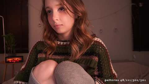 Media: Video of a young girl with wavy, light brown hair, wearing a green, patterned sweater, sitting on a bed in a dimly lit room with a fireplace and string lights.