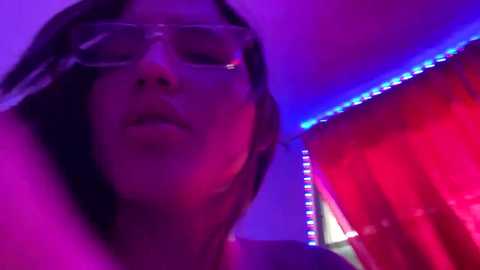 Media: A video of a person with light skin, wearing glasses and a hooded top, under purple and red LED lighting, with blurred background.