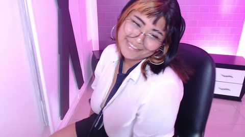 Media: Video of a smiling, middle-aged Asian woman with glasses and short black hair, wearing a white lab coat and black shirt, sitting in a modern office with pink lighting and minimalist decor.