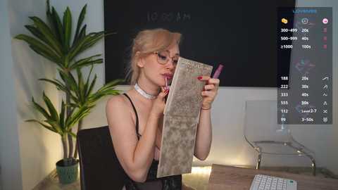 Media: A video of a blonde woman in glasses and a choker, reading a book while holding a pen, in a modern office with a green plant, blackboard, and white chair.