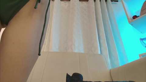 Video of a woman in a green top, partially undressing, with a white curtain in the background, revealing a glimpse of her bare back and light skin tone.